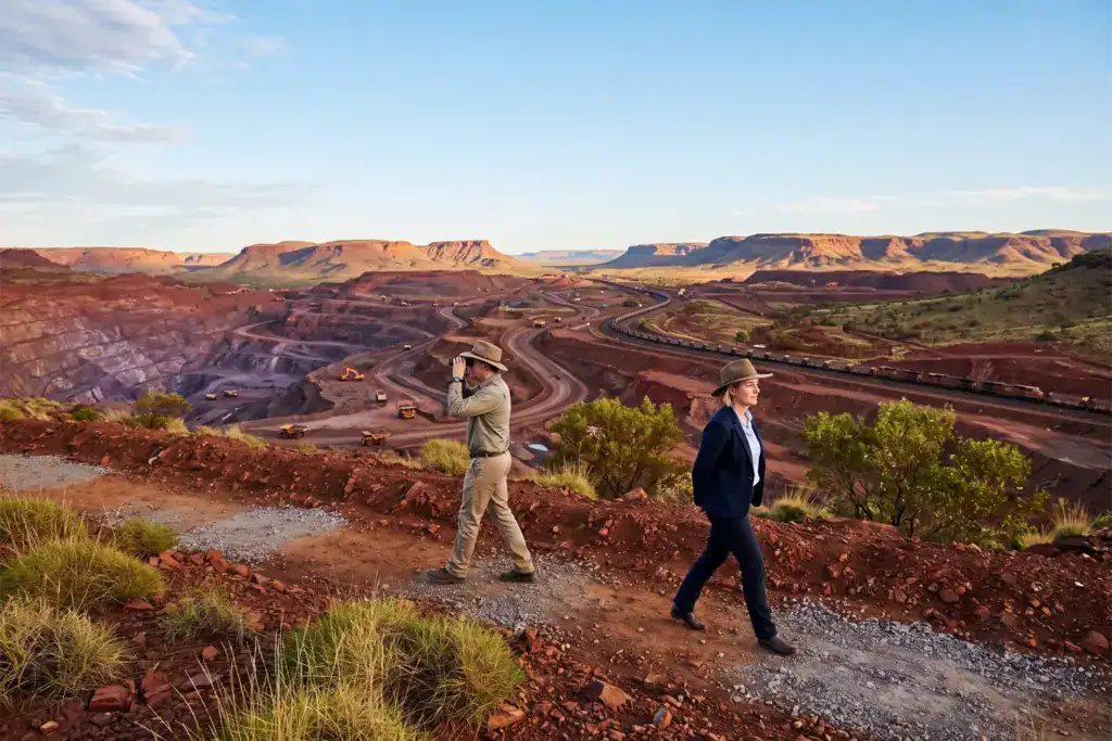 A man and a woman walk on separate paths above a large Australian mining landscape beneath bright skies.