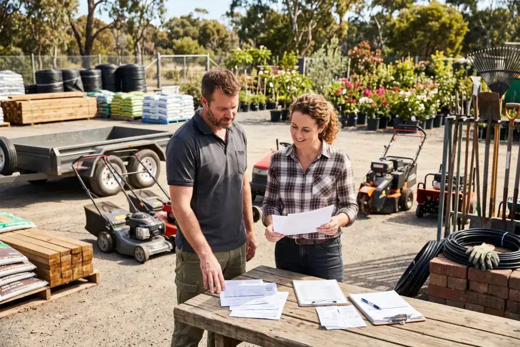 A man and a woman stand beside landscaping equipment while they examine printed financial papers on a timber table. The image represents small businesses making the most of federal budget australia opportunities.